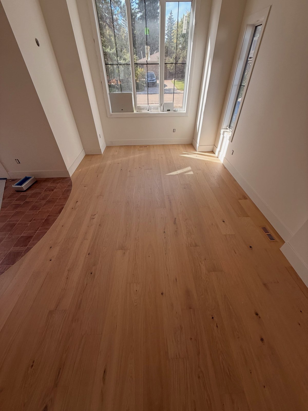 Warm tan plank flooring beneath tall windows, with a curved transition to a reddish patterned floor on the left.