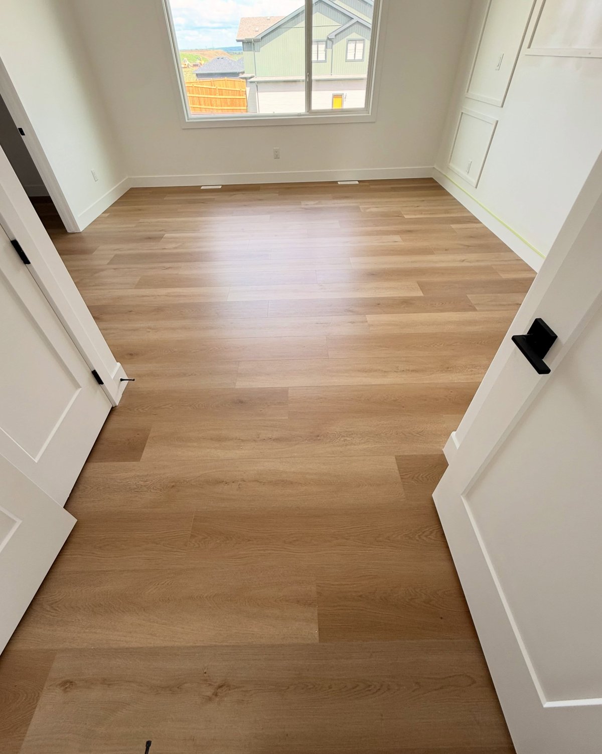 Warm tan plank flooring with staggered joints viewed through open double doors into an empty room with a window.