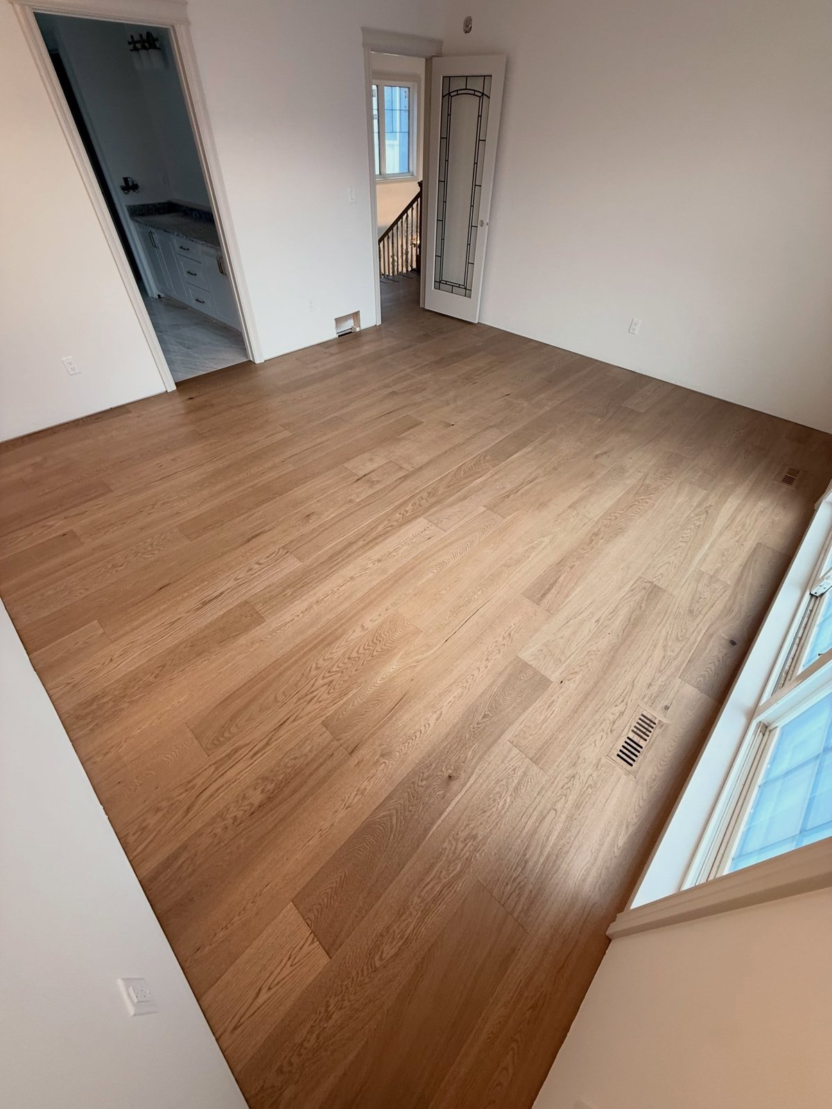 Tan wood-grain plank flooring with staggered joints in an empty room, with a floor register beside the right-hand window.